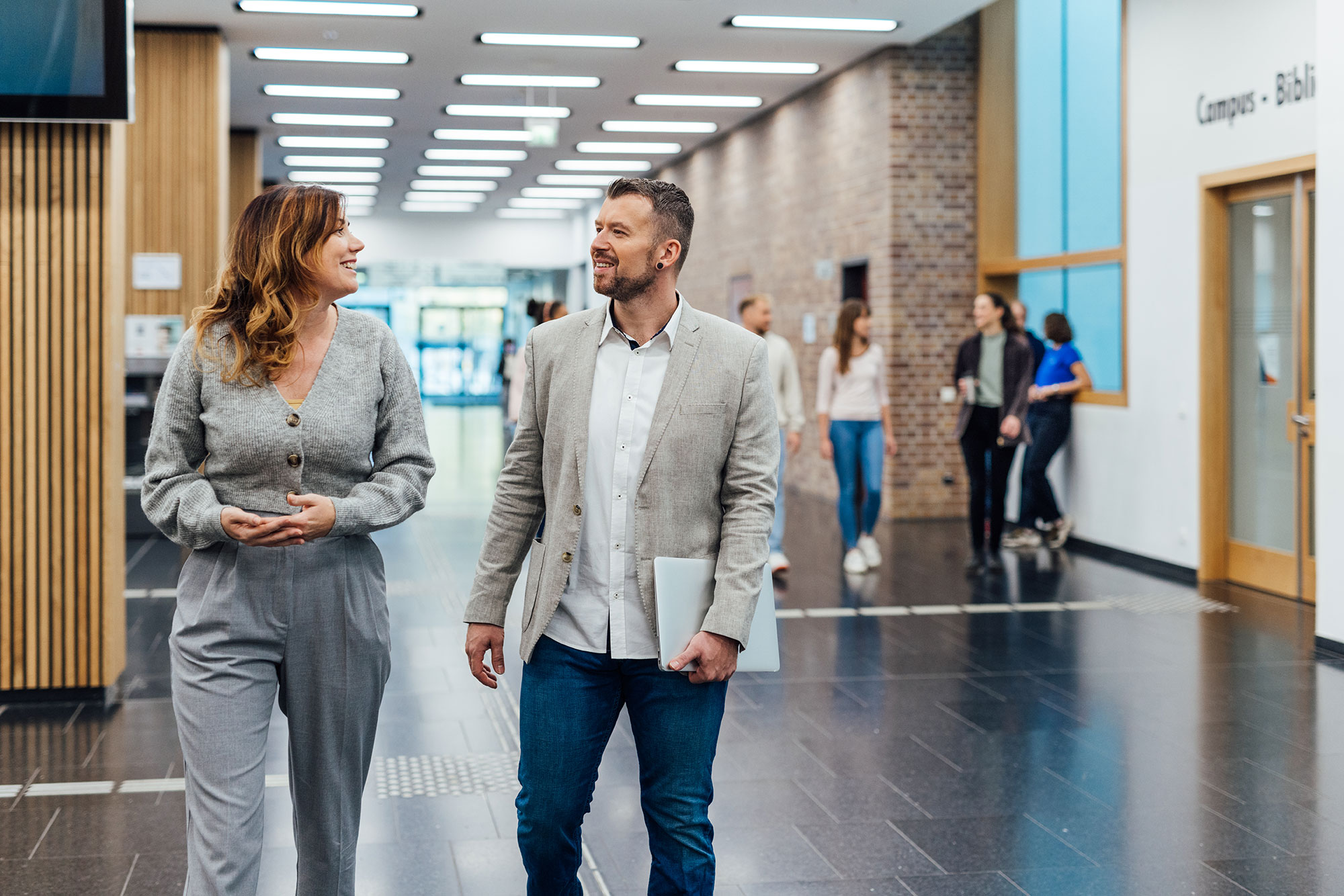 Ein Mann und eine Frau gehen einen langen Flur in einem Hochschulgebäude entlang und unterhalten sich miteinander.