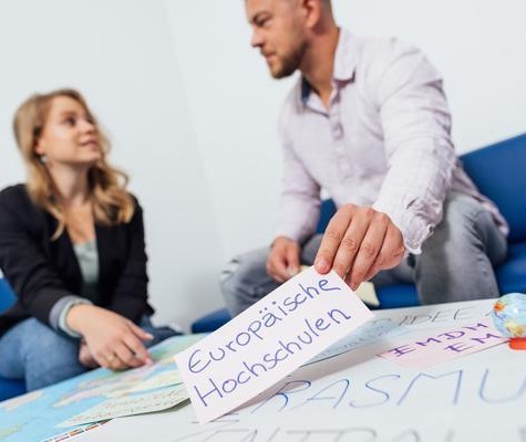 Zwei Personen sitzen auf einem Sofa und planen ein Erasmus+ Projekt. Im Vordergrund hält eine Person eine Karte mit der Aufschrift „Europäische Hochschulen“ über einer Arbeitsfläche mit Notizen und Materialien.