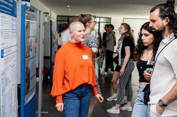 Mehrere Personen stehen in einem Innenraum vor wissenschaftlichen Postern und kommen miteinander ins Gespräch. Eine Person in orangefarbener Bluse erklärt Inhalte, während andere aufmerksam zuhören; im Hintergrund sind weitere Poster und Besucher zu sehen.