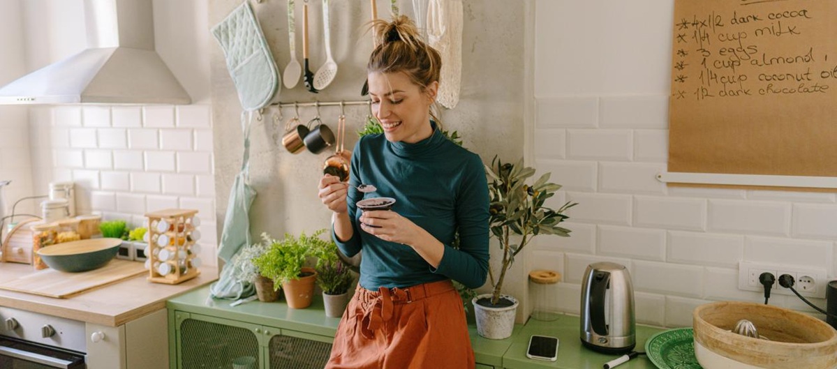 Junge Frau steht in der Küche mit einem Joghurt in der Hand.